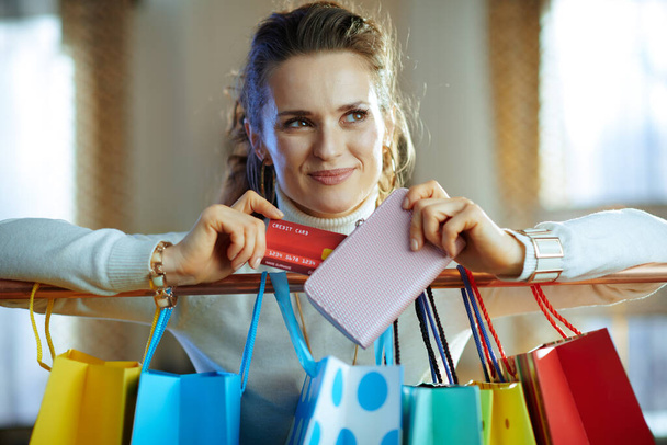 happy stylish 40 years old woman in white sweater and skirt near colorful shopping bags hanging on copper clothes rail taking out credit card from wallet. - Foto, Imagem