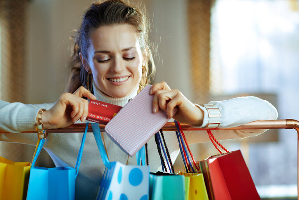 happy modern woman in white sweater and skirt near colorful shopping bags hanging on copper clothes rail taking out credit card from wallet. - Foto, imagen