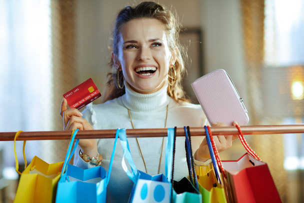 happy stylish female in white sweater and skirt with credit card and wallet near colorful shopping bags hanging on copper clothes rail. - Foto, Bild
