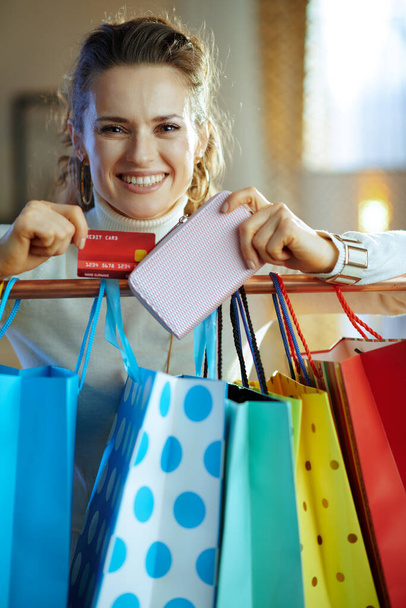 mujer elegante feliz en suéter blanco y falda cerca de coloridas bolsas de compras colgando en el carril de cobre ropa sacar tarjeta de crédito de la cartera. - Foto, imagen