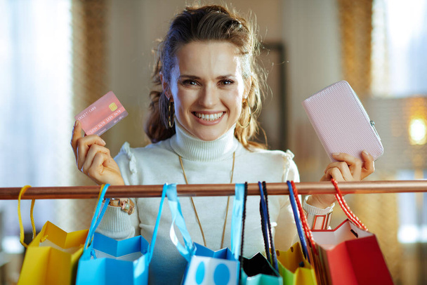 smiling trendy middle age woman in white sweater and skirt with credit card and wallet near colorful shopping bags hanging on copper clothes rail. - Valokuva, kuva