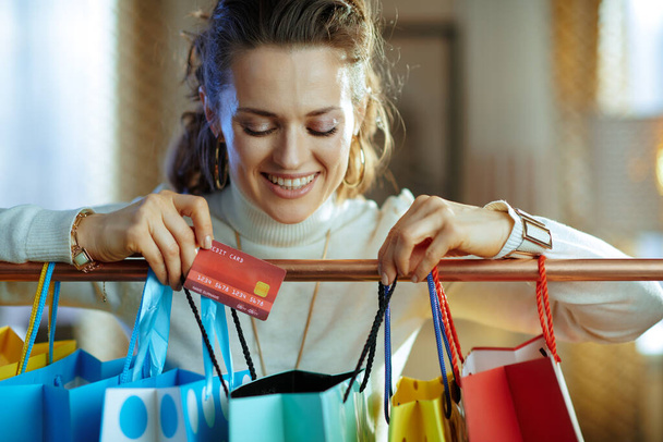 smiling modern 40 years old woman in white sweater and skirt looking in shopping bag hanging on copper clothes rail with credit card. - Photo, Image
