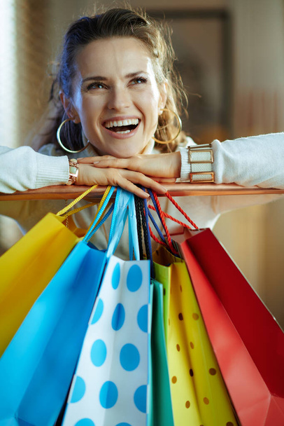 smiling elegant 40 years old woman in white sweater and skirt looking into the distance near colorful shopping bags hanging on copper clothes rail. - 写真・画像