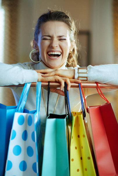 cheerful elegant middle age woman in white sweater and skirt near colorful shopping bags hanging on copper clothes rail. - Foto, imagen