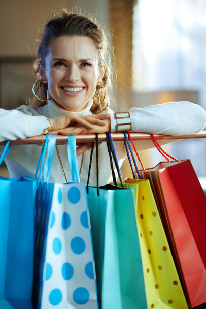 happy trendy 40 years old woman in white sweater and skirt near colorful shopping bags hanging on copper clothes rail. - Foto, imagen
