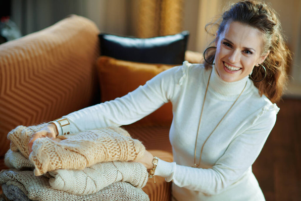 feliz mujer de mediana edad de moda en suéter blanco y falda en el hogar moderno en el día de invierno soleado sentado cerca de los suéteres plegables sofá para el almacenamiento. - Foto, imagen