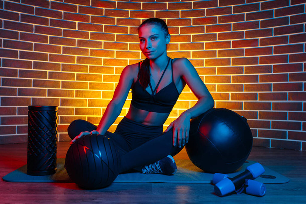 Fit young brunette woman posing with medicine balls and foam fitness roller on mat in gym in neon lights. - Foto, imagen