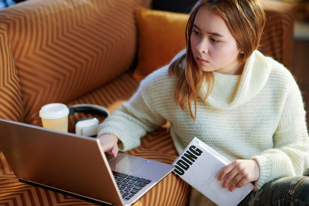 concerned modern teen girl with red hair in white sweater with laptop and book learning coding in the modern living room in sunny winter day. - Fotografie, Obrázek