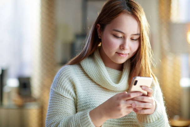happy modern teen girl with red hair in white sweater at modern home in sunny winter day browsing social media on a smartphone. - Photo, image