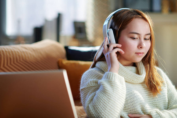 modern teen girl with red hair in white sweater listening to the music with headphones while sitting near couch and laptop in the modern living room in sunny winter day. - Foto, imagen