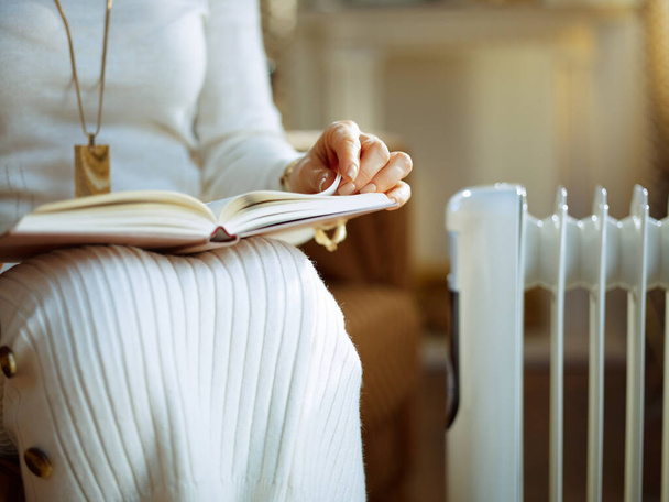 Closeup on middle age housewife in white sweater and skirt at modern home in sunny winter day sitting on couch near white electric oil radiator and reading book. - Fotó, kép