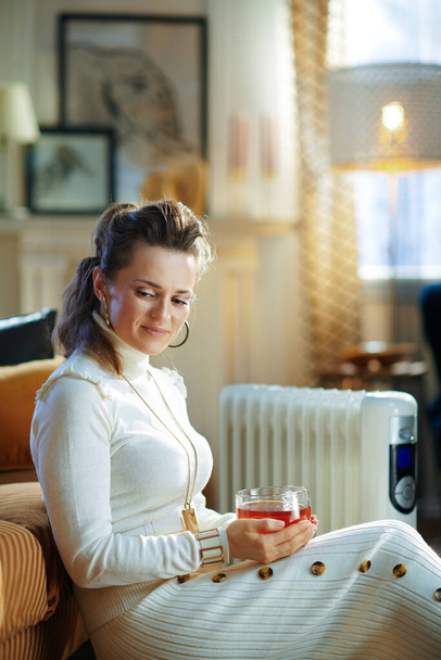 töprengő trendi nő fehér pulóverben és szoknya a modern házban napfényes téli nap közelében ül kanapé fehér elektromos olaj radiátor és gazdaság csésze forró tea. - Fotó, kép
