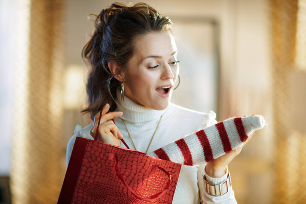 yllättynyt moderni keski-ikäinen nainen valkoisessa villapaita ja hame punainen ostoskassi ottaa ostettu villapaita moderni olohuone aurinkoinen talvipäivä. - Valokuva, kuva