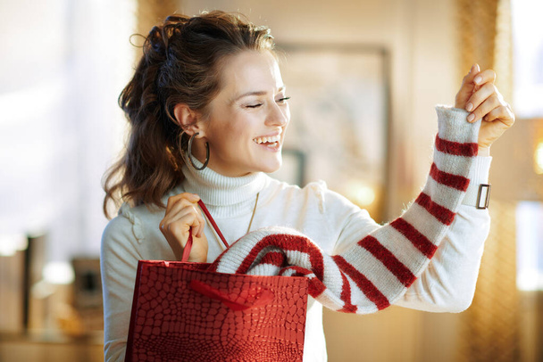 smiling elegant middle age housewife in white sweater and skirt with red shopping bag checking purchased sweater in the modern house in sunny winter day. - Zdjęcie, obraz