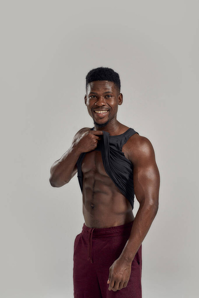 Young muscular african american man smiling at camera, lifting black shirt, showing his abs while posing isolated over grey background. Sports, workout, bodybuilding concept - Фото, изображение