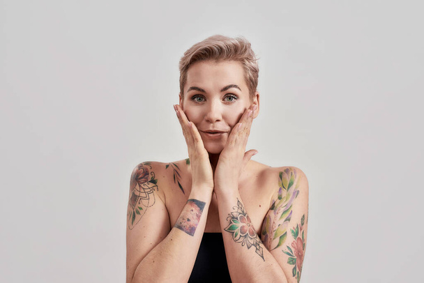 Portrait of half naked tattooed woman with short hair smiling at camera, touching her face isolated over light background - Фото, изображение
