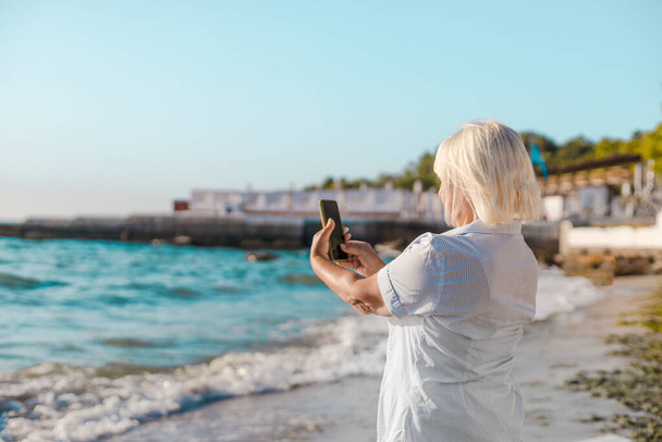 Happy woman on vacation photographing with a camera on the beach of the sea by smartphone, enjoys leisure and summer vacation - Foto, immagini