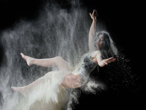 schöne junge kaukasische Frau in einem schwarzen Body mit einer sportlichen Figur schwebt auf einem schwarzen Hintergrund in einem Spray aus weißem Mehl, kreatives Foto, Schwebeeffekt - Foto, Bild