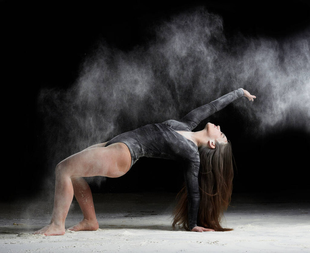 young beautiful caucasian woman with long hair is dressed in a sports black bodysuit stands in a bent pose and throws white flour, black background, gymnastic exercises - Foto, Imagen