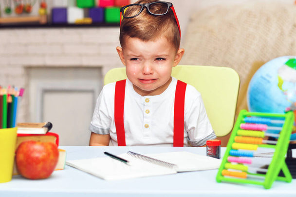Un niño en edad preescolar se sienta en un escritorio con una camiseta blanca y tirantes rojos y llorando. - Foto, Imagen