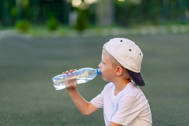 portrét blonďatého chlapce ve sportovní uniformě sedícího na zeleném trávníku na fotbalovém hřišti a pijícího vodu z láhve, sportovní sekce. Školení dětí, dětský volný čas. - Fotografie, Obrázek