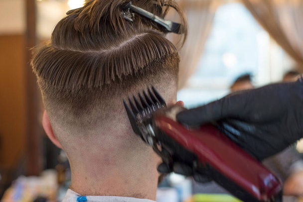 Ein Friseur schneidet einen bärtigen jungen Mann mit einem Haarschneider und kämmt ihm die Haare auf dem Kopf. Arbeit des Friseurmeisters in einem Friseursalon. Die Schönheit der Männer, lineare Trimmfrisuren. Phasen des Haarschnitts auf dem Kopf bei Männern, die Vorbereitung der Haare für - Foto, Bild