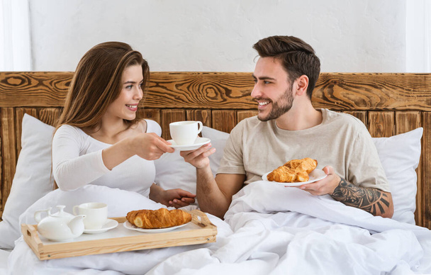 Romantic breakfast of couple in bed. Smiling attractive young woman holding tray and giving a cup of coffee to man - Photo, image