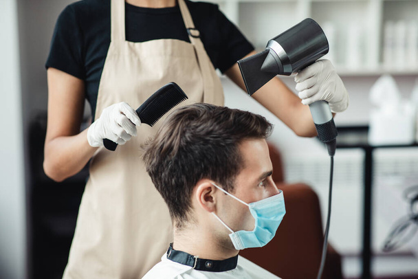 Jeune homme en masque médical obtenir coiffure au salon de coiffure, vue de côté - Photo, image