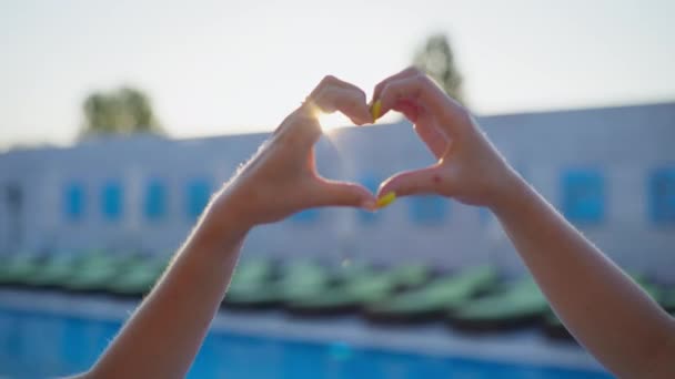 fim de semana de verão, belas mãos femininas mostram um coração no fundo de raios de sol e espreguiçadeiras com uma piscina, close-up - Filmagem, Vídeo