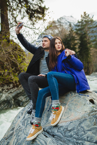 young hipster beautiful couple in love holding smartphone, taking pictures, sitting on a rock at river in winter forest, jeans, warm coat, sneakers, smiling, having fun, traveling, hiking, vacation - Fotografie, Obrázek