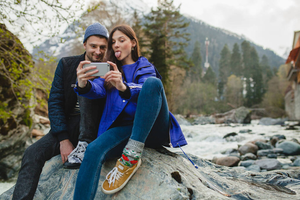 young hipster beautiful couple in love holding smartphone, taking pictures, sitting on a rock at river in winter forest, jeans, warm coat, sneakers, smiling, having fun, traveling, hiking, vacation - Фото, изображение