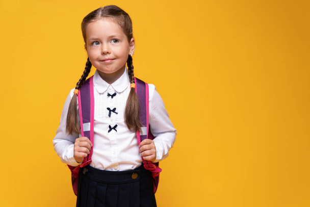 smiling little cute school girl in uniform with school backpack, isolated on yellow background - Фото, изображение