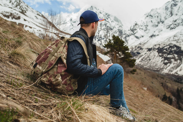 young hipster man sitting in mountains, swag outfit, winter vacation, trendy style, having fun, jeans, backpack, hiking, jacket, cap, sneakers - Fotoğraf, Görsel