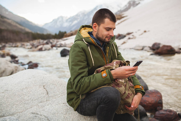 young hipster man using smartphone, wild nature, winter vacation, hiking, traveling, backpacker, warm clothes - Photo, Image