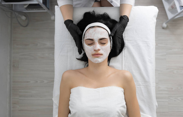 A kozmetikus enzimatikus hámlasztást alkalmaz a nő arcán a fürdőben. Fehér maszkot visel ecsettel. Kozmetikai eljárás egy modern szépségszalonban. portré nézet - Fotó, kép