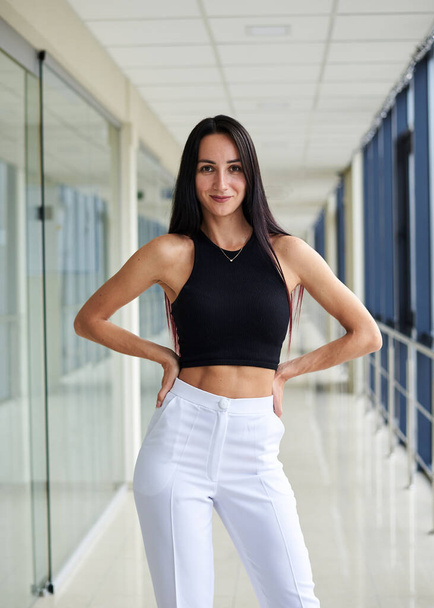 Young brunette woman, wearing white pants and black top, standing in light passageway with huge windows, posing with arms on her waist. Businesswoman on a break. Female portrait photography. - Fotografie, Obrázek