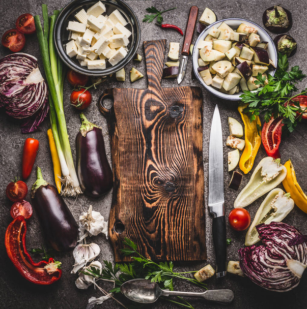 Various vegetables cooking ingredients around wooden cutting board with knife on rustic background with bowls. Vegetarian cooking preparation for tasty dishes, top view. Healthy eating concept - Foto, Imagen