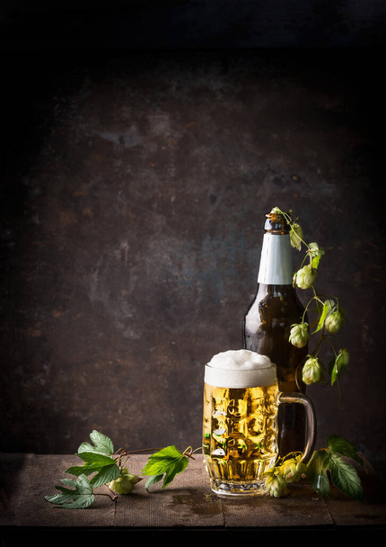 Glass bottles and mug of beer with cap of foam and hops on table at dark rustic background, front view, Still life - Fotoğraf, Görsel