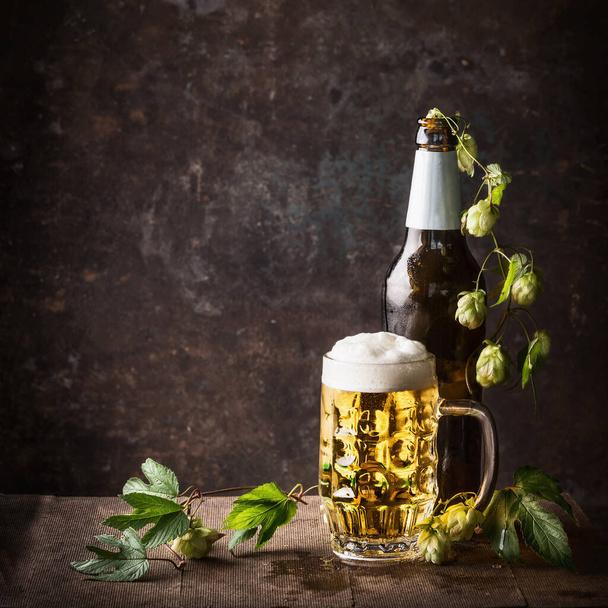 Glass bottles and mug of beer with cap of foam and hops on table at dark rustic background, front view, Still life , close up - Photo, Image