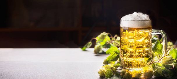 Mug of beer with foam on table with hops at dark background , front view, banner - Fotó, kép