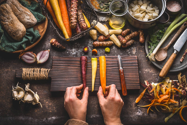 Healthy vegetables cooking and eating concept. Female woman hands holding colorful carrots on kitchen table background with vegetables peeler, knife, cutting board and cooking pot, top view - Foto, Imagem