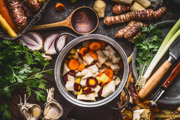Organic farm vegetables cooking and eating. Cooking pot with diced colorful root vegetables on rustic kitchen tables background with spoon and ingredients. Healthy clean food concept - Foto, imagen
