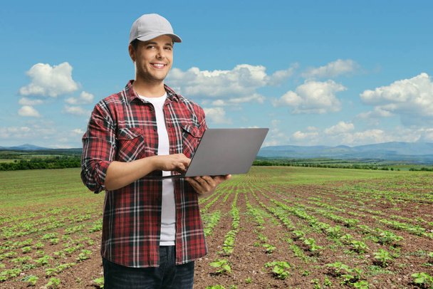 Young man in a checkered shirt holding a laptop computer and looking at the camera on a field with plants - Foto, afbeelding