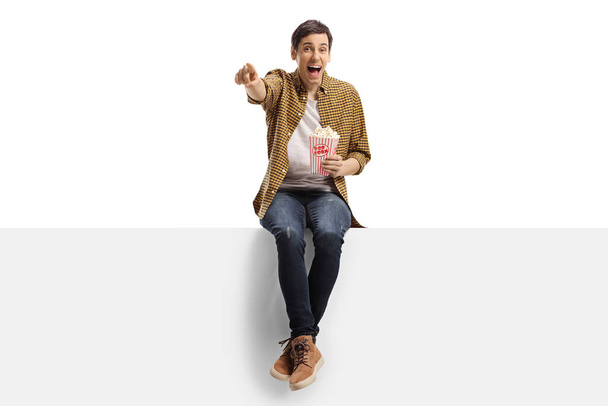 Νεαρός άνδρας γελάει και δείχνει ένα πάνελ και τρώει ποπ κορν απομονωμένο σε λευκό φόντο - Φωτογραφία, εικόνα