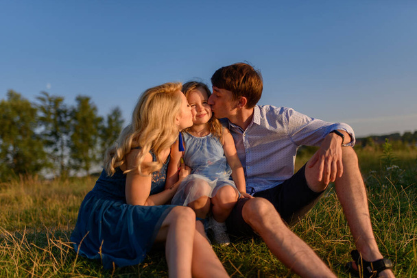 Papá, mamá e hija están sentadas juntas en la hierba. Los padres besan a su hija en las mejillas. - Foto, imagen