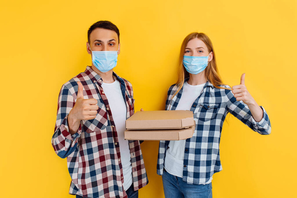 man and a woman in plaid shirts and medical protective masks on their faces, holding pizza boxes while giving a thumbs-up on an isolated yellow background - 写真・画像