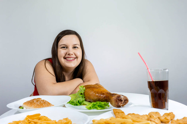 fat girl sits at a table and eats dinner in which fast food: French fries, burger, chicken leg, chips. - Foto, Imagen