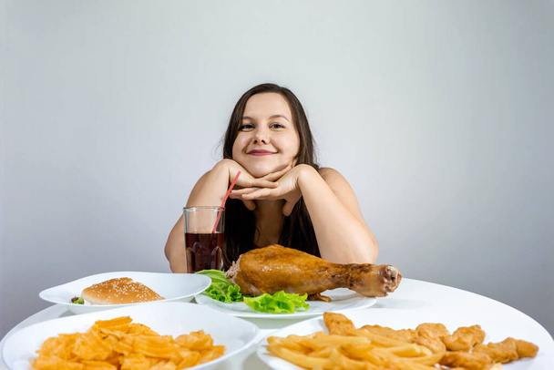 woman sits at a table and eats dinner in which fast food: French fries, burger, chicken leg, chips. - Foto, Imagen