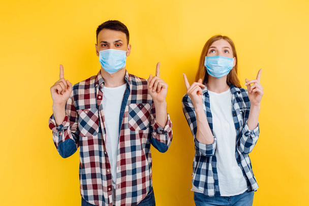man and a woman in checked shirts and medical protective masks on their faces, pointing to a copy of the space on an isolated yellow background - Фото, изображение