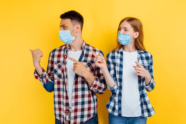 man and a woman in checked shirts and medical protective masks on their faces, pointing their fingers at a copy of the space on an isolated yellow background - Photo, Image